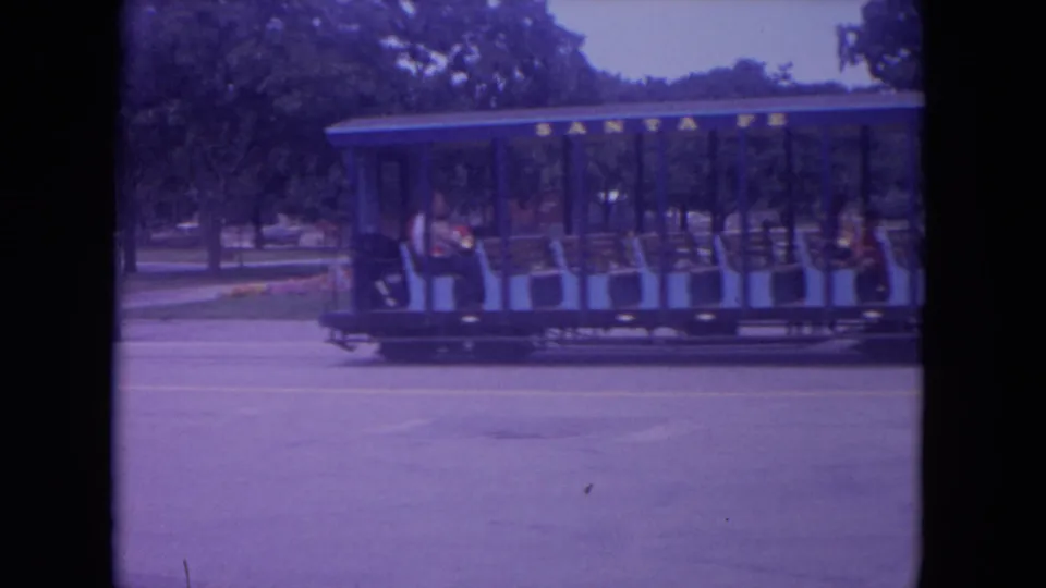 A blue and green open-sided train car passes through a park-like area in the 1972 Colby, Wisconsin home-movie footage.