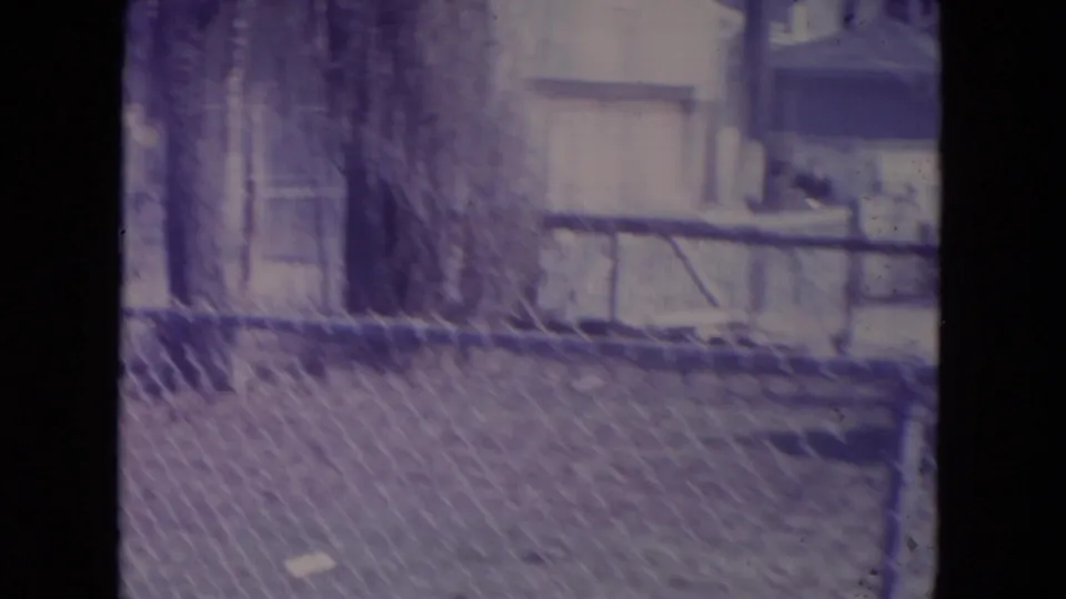 A 1972 home-movie view across a chain-link fence toward a tree, garage, and residential yard in Racine.