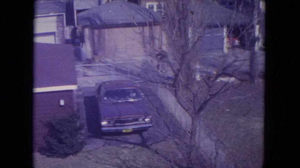 Residential driveway scene with an older sedan and bare tree in Racine home-movie footage.