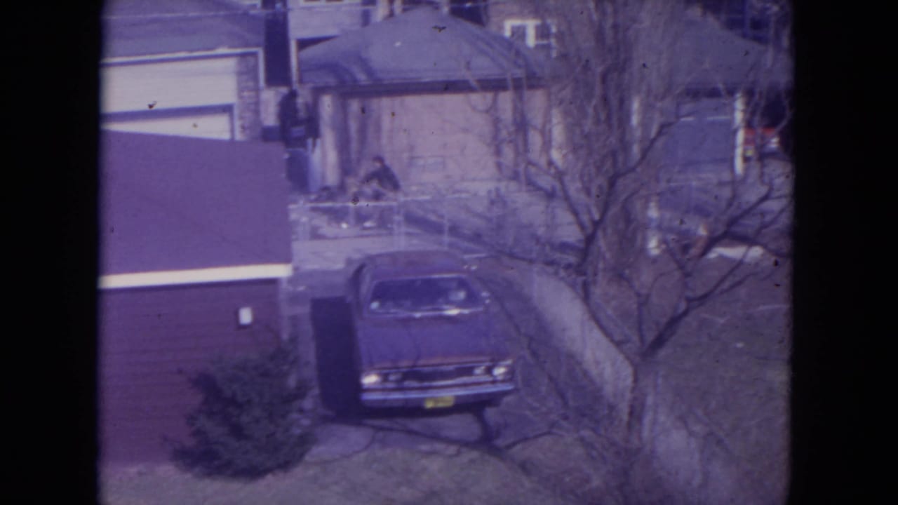Elevated view of a parked sedan beside houses, garages, bare trees, and a cyclist in the background.