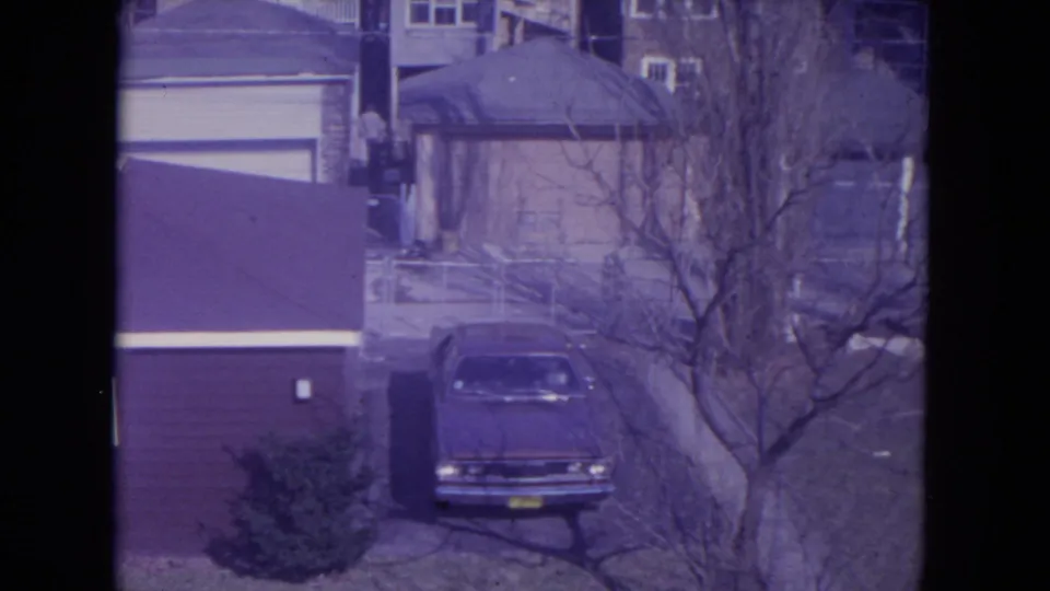 Racine, Wisconsin 1972 home-movie frame of a parked sedan near house siding, a garage, bare trees, and a small residential lawn.