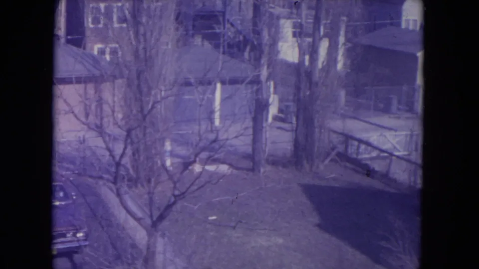 1972 Racine neighborhood frame showing bare trees, neighboring houses, porch railings, lawns, and the edge of a parked car at lower left.