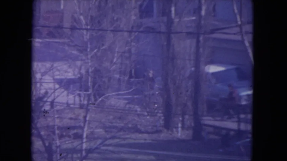 Racine 1972 home-movie frame showing bare trees, a residential yard, a house, and a car with visible 8mm grain.