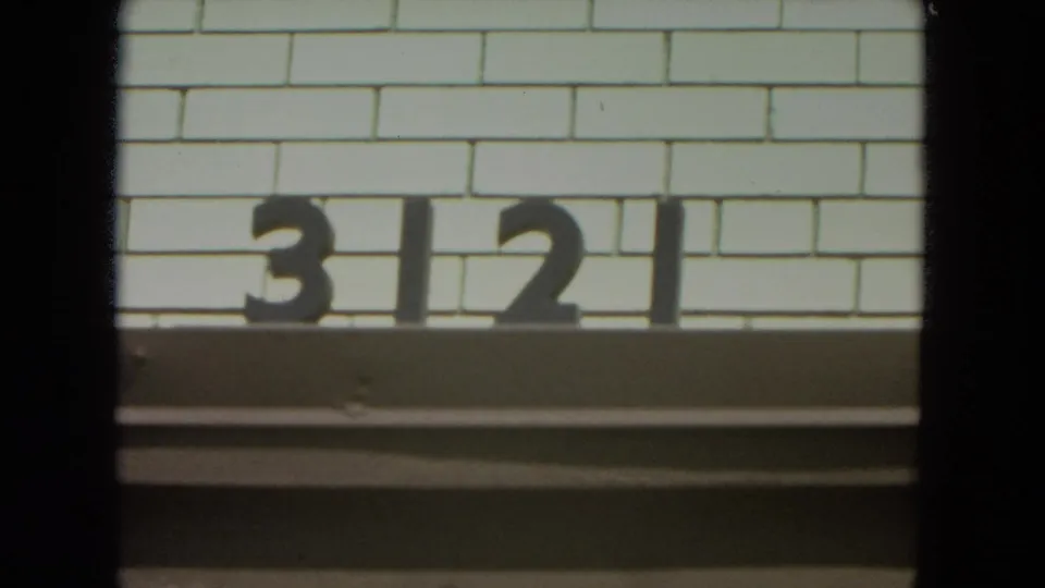 1972 Racine home-movie frame showing black 3121 numerals on a pale brick building facade, partially bordered by dark film or window edges.