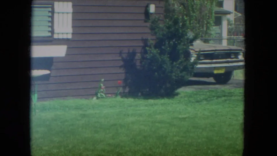 1972 Racine residential exterior with green lawn, brown siding, a shrub near the wall, and part of an older car in the driveway.