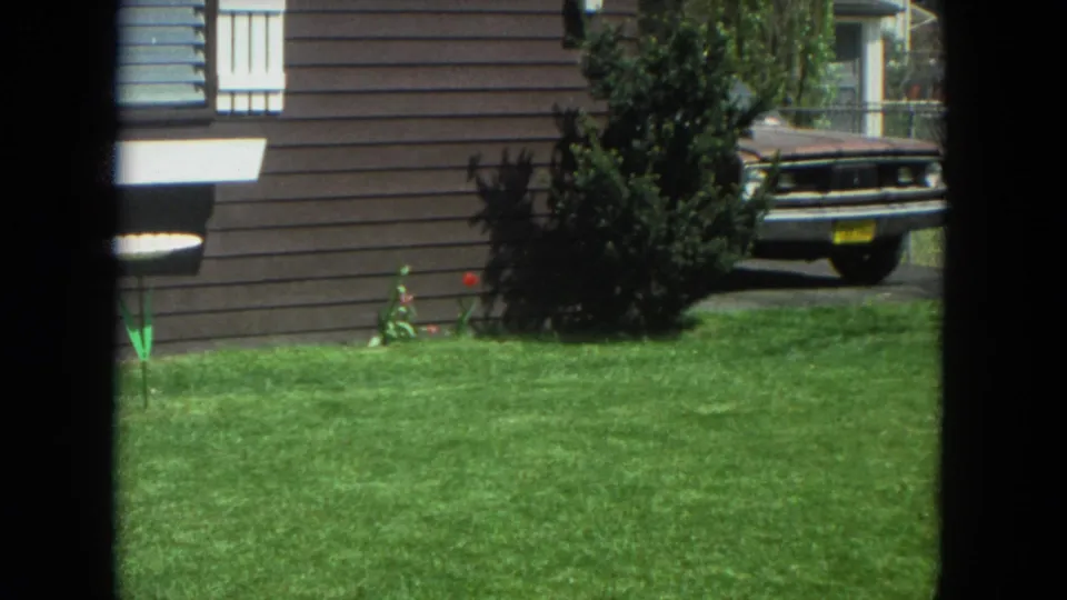 1972 Racine home-movie view of a suburban front yard with brown siding, shrubbery, flowers, and an older parked car near the driveway.