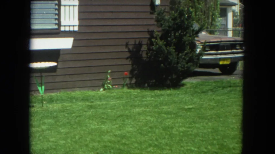 1972 Racine residential exterior showing a green lawn, brown house siding, shrubs, flowers, and a car partly framed at the driveway.