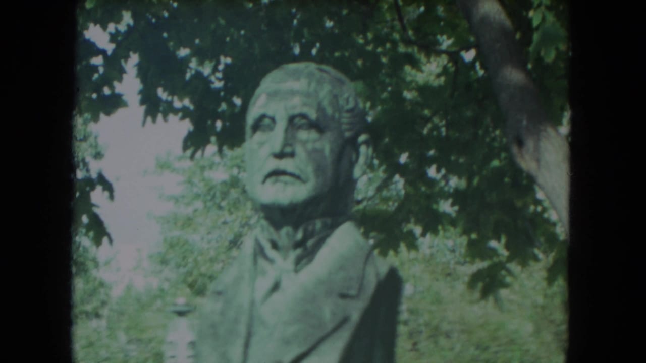Bronze bust of a man outdoors beneath leafy trees.