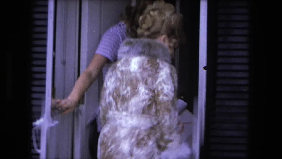 In Fort Wayne home-movie footage from 1973, a woman in a patterned coat exits while another woman stands in the doorway of a suburban house.