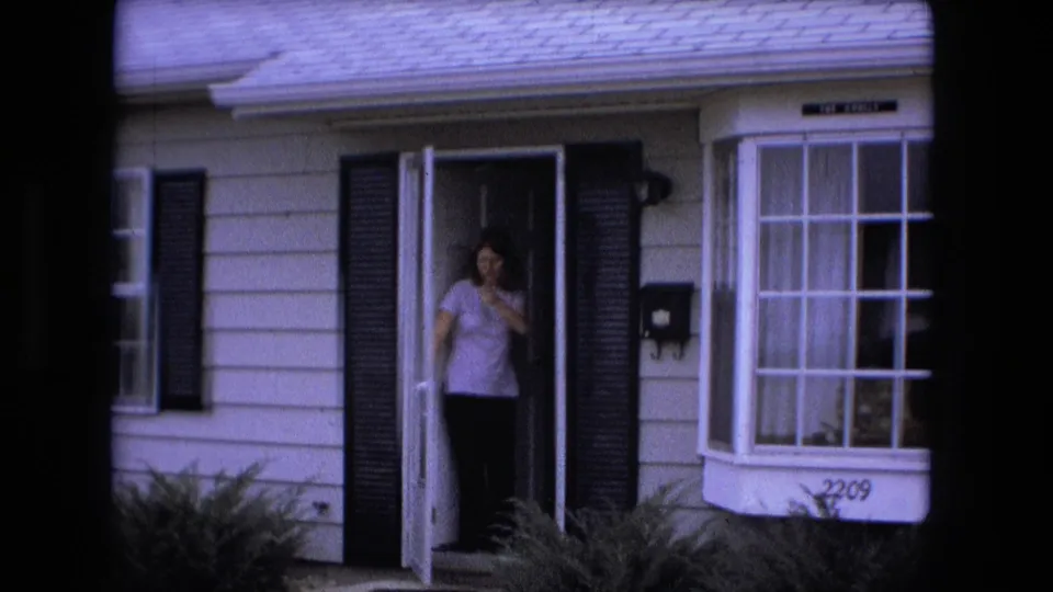 A woman waves from the doorway of a Fort Wayne house, with black shutters, white siding, and the house number visible below the bay window.