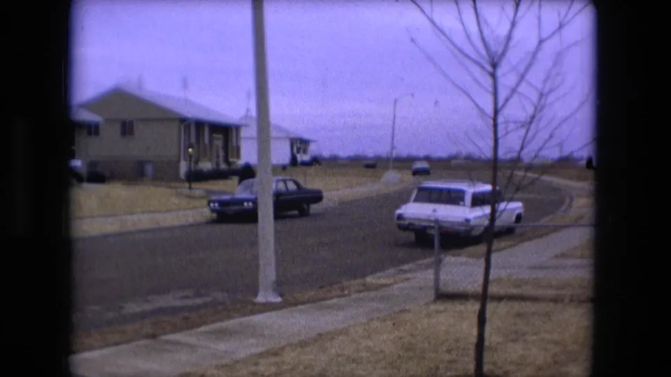 A Fort Wayne suburban street view shows older cars, sidewalks, lawns, bare trees, and low residential buildings in 1973 home-movie footage.