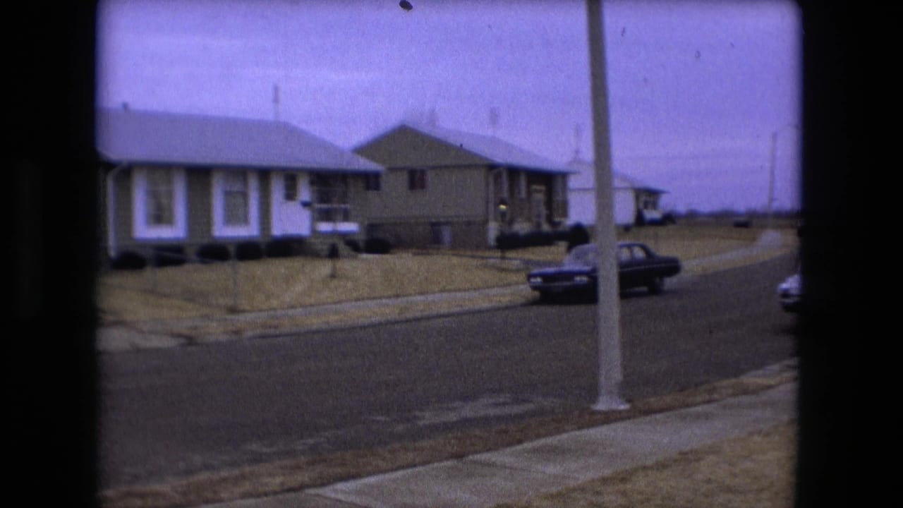 Suburban street corner with houses, a dark car, sidewalks, and pale winter or fall lawns.