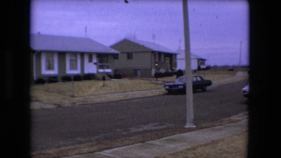 Fort Wayne suburban block in 1973 with green-and-white houses, a dark sedan near the curb, sidewalks, and leafless trees.