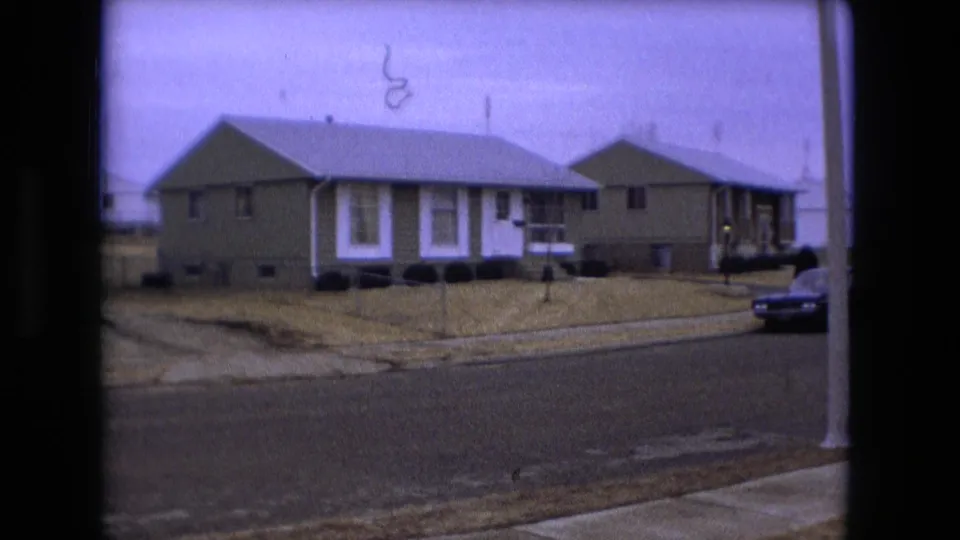 1973 Fort Wayne residential view with low houses, dormant lawns, a streetlight pole, bare branches, and a dark car at frame edge.