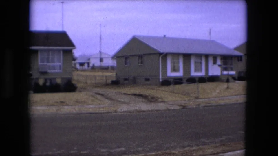 Overcast 1973 Fort Wayne street corner with single-story homes, a neighboring porch or sunroom, bare lawns, and a quiet road.