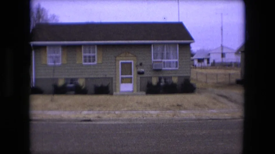 Closer 1973 Fort Wayne frame of a green suburban house with shutters, screen door, front windows, and dry lawn.