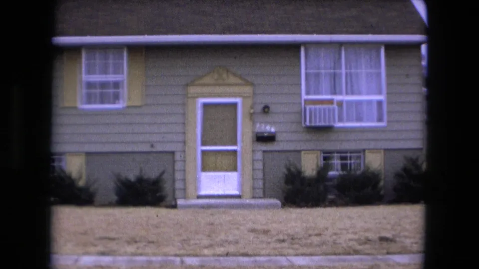 Tight 1973 Fort Wayne frame showing the green house facade, white-trimmed door, lower windows, shrubs, and window air conditioner.