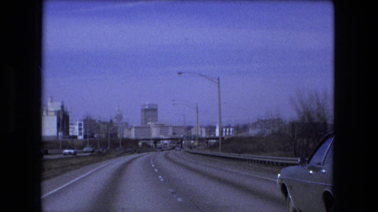 Forward view down a multi-lane highway with guardrails, streetlights, distant buildings, and part of a car at right.