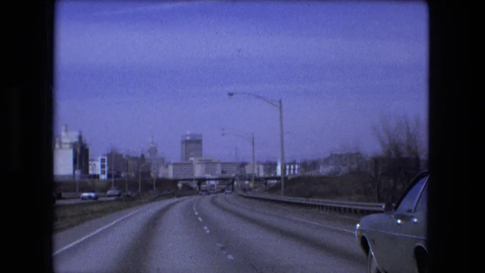 1973 Fort Wayne highway approach seen from a moving vehicle, with dashed lanes, guardrails, streetlights, distant structures, and a car beside the camera.