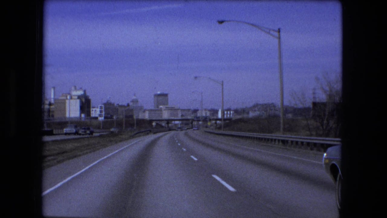 1973 Fort Wayne road view from a moving car, showing a curving multi-lane highway, overpass ahead, tall streetlights, and city-edge buildings.