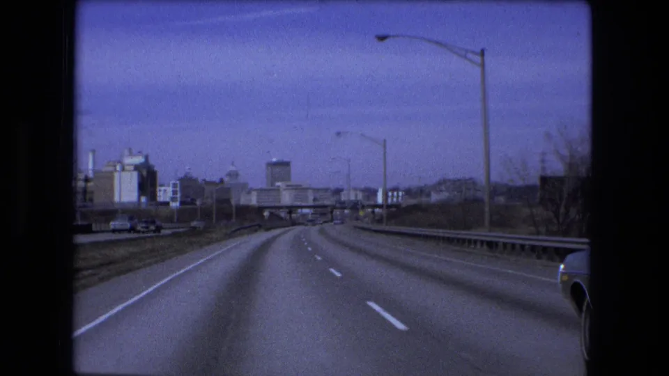 1973 Fort Wayne road view from a moving car, showing a curving multi-lane highway, overpass ahead, tall streetlights, and city-edge buildings.
