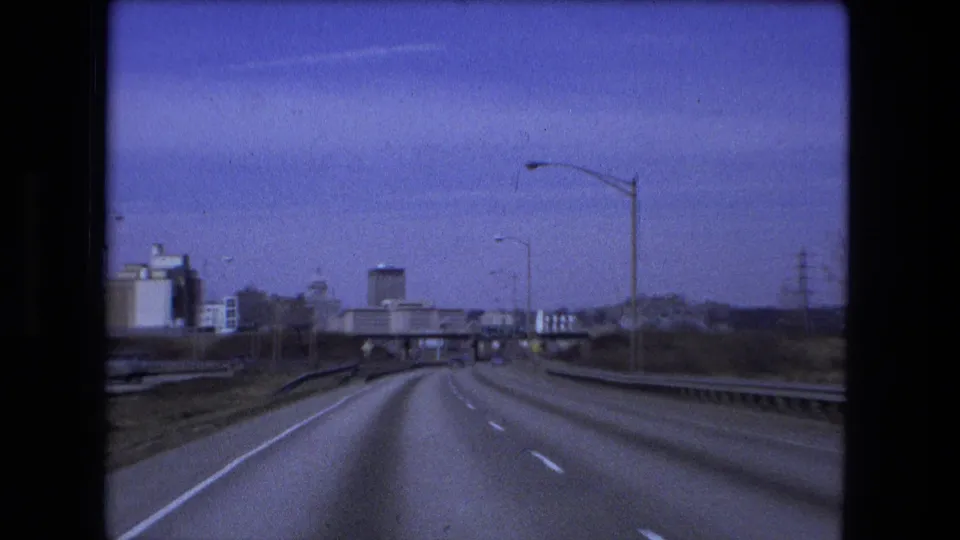 1973 Fort Wayne highway footage shows a forward road perspective with wide lanes, roadside guardrails, overhead approach, and distant urban structures.
