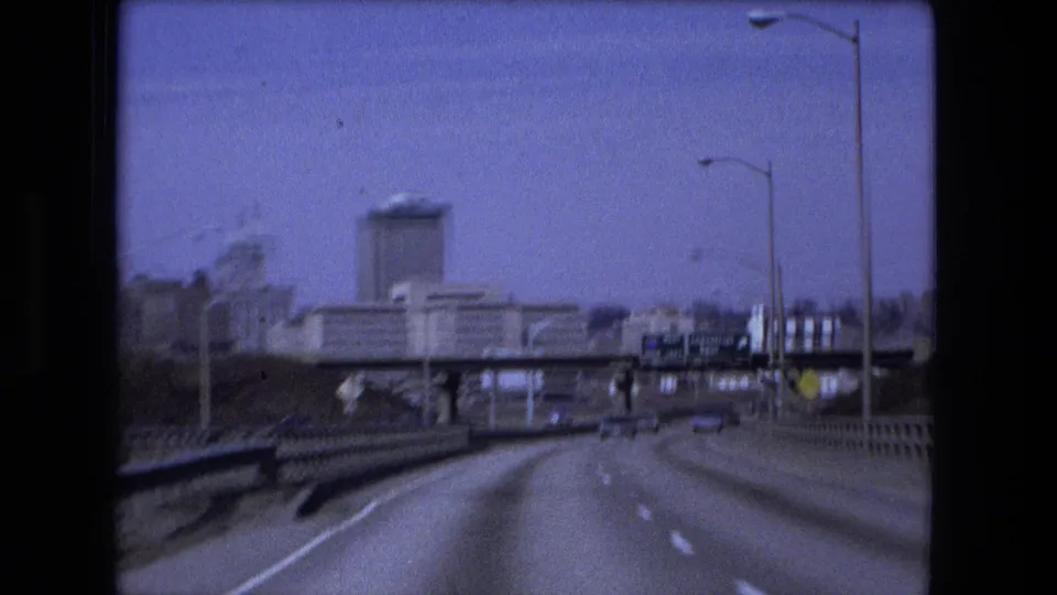 1973 Fort Wayne driving view nearing an overpass, with a prominent tower-like structure, roadside lights, guardrails, and sparse traffic in the distance.