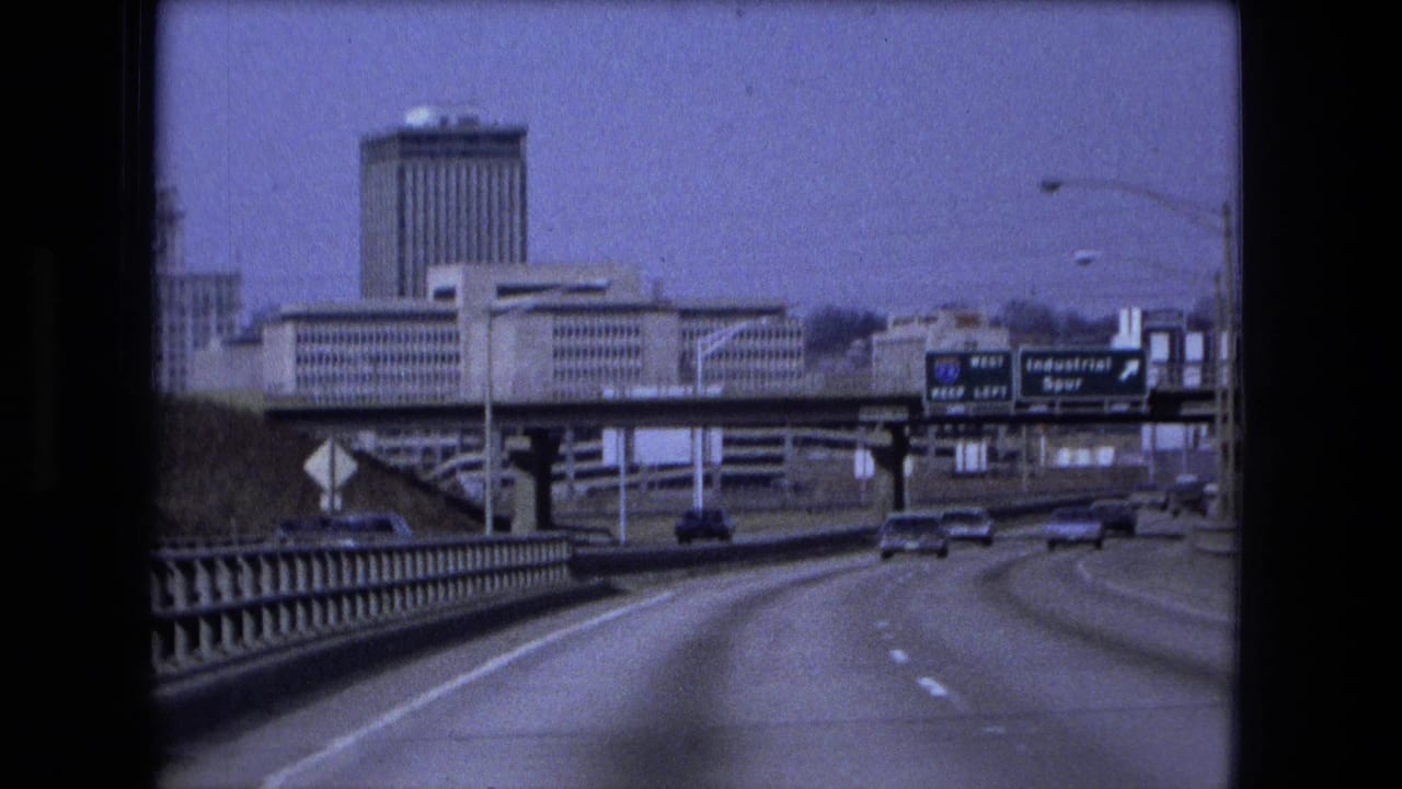 Roadway approach with downtown buildings, bridge railing, overhead signs, and cars ahead.