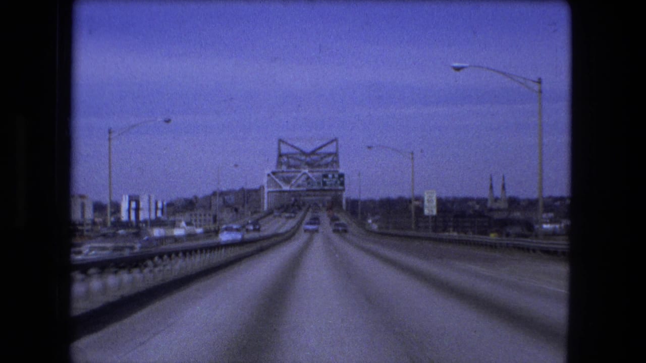 1973 Fort Wayne bridge approach seen from a moving vehicle, with a truss span ahead, cars on the roadway, railings, and distant city structures.