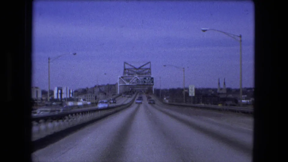 1973 Fort Wayne bridge approach seen from a moving vehicle, with a truss span ahead, cars on the roadway, railings, and distant city structures.