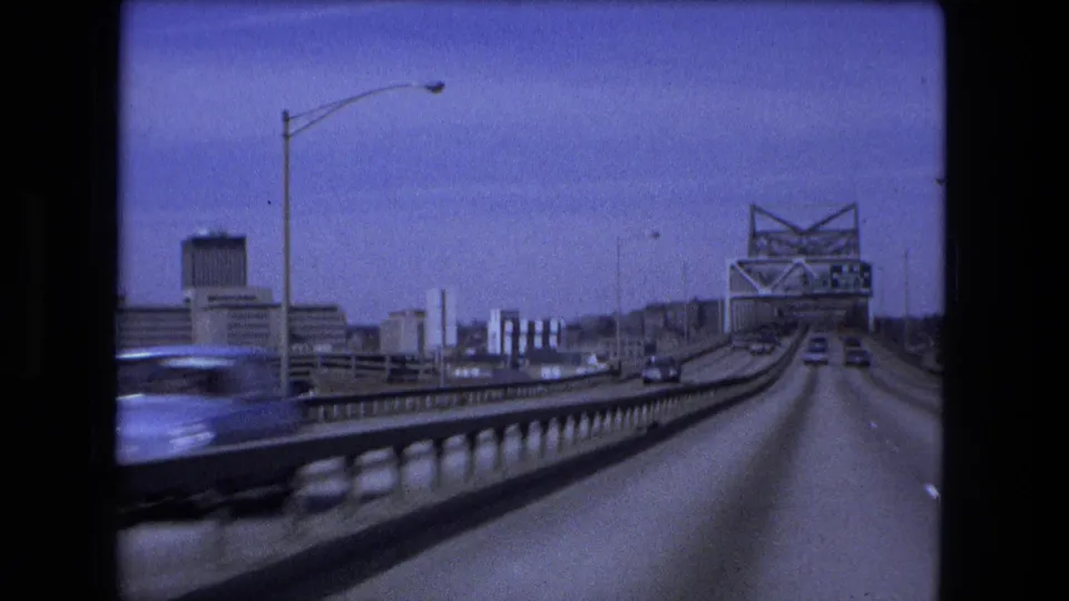 1973 Fort Wayne bridge crossing from a vehicle, with a blurred passing car, roadway lanes, bridge railings, and downtown buildings in the distance.