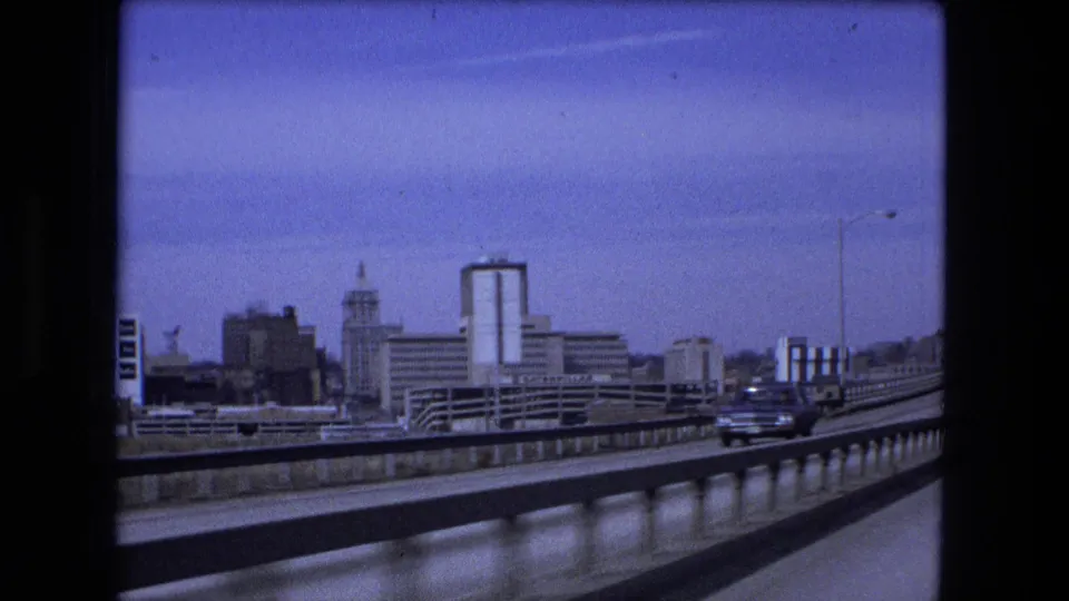 1973 Fort Wayne skyline viewed from a bridge roadway, showing railings, cars, streetlights, and downtown mid-rise buildings under clear sky.