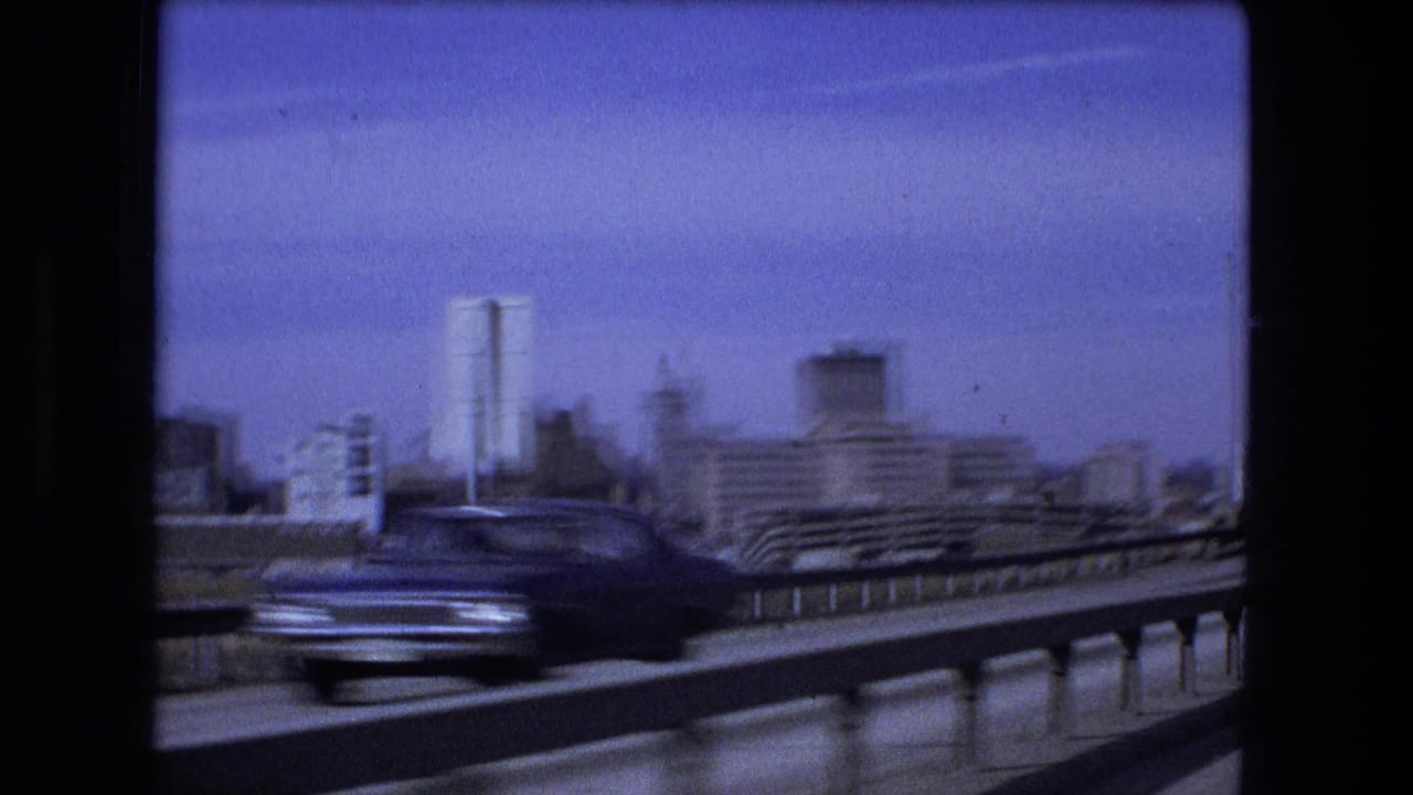 Moving highway view with a blurred car, guardrail, and distant city buildings.