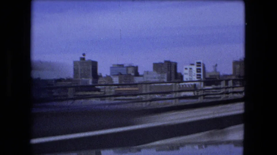 A moving 1973 Fort Wayne roadway view shows low-rise city buildings, a barrier, and open blue sky.