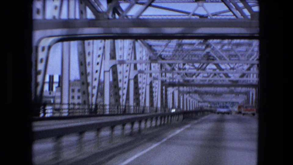 Vehicle enters a steel truss bridge in Fort Wayne, with roadway lanes, bridge beams, and distant traffic visible in the 1973 8mm frame.