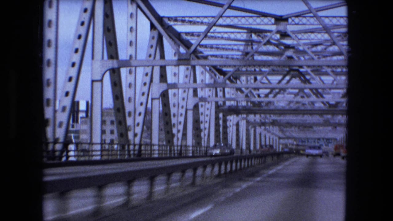 Forward view along a steel truss bridge roadway with cars in the distance.