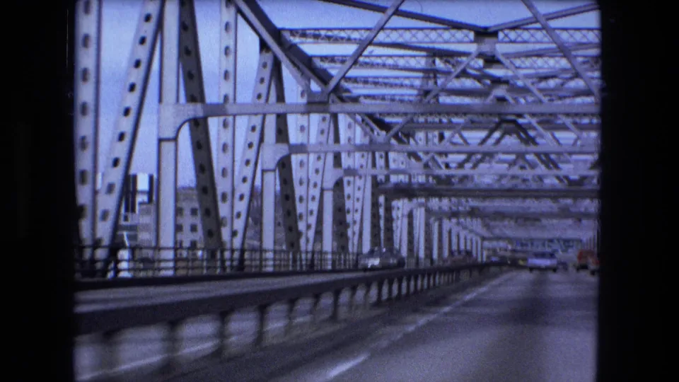 1973 Fort Wayne home-movie frame looking forward across a steel truss bridge, with roadway lanes, guardrails, and distant vehicles visible through the metal framework.