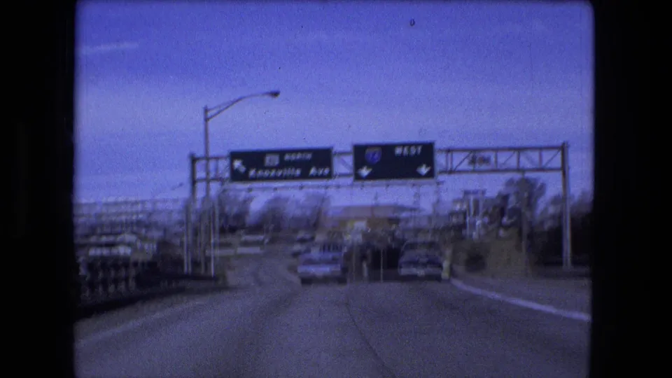 Forward road view in Fort Wayne with overhead highway signs, multiple cars, bridge railings, and a broad roadway leading away from the bridge crossing.