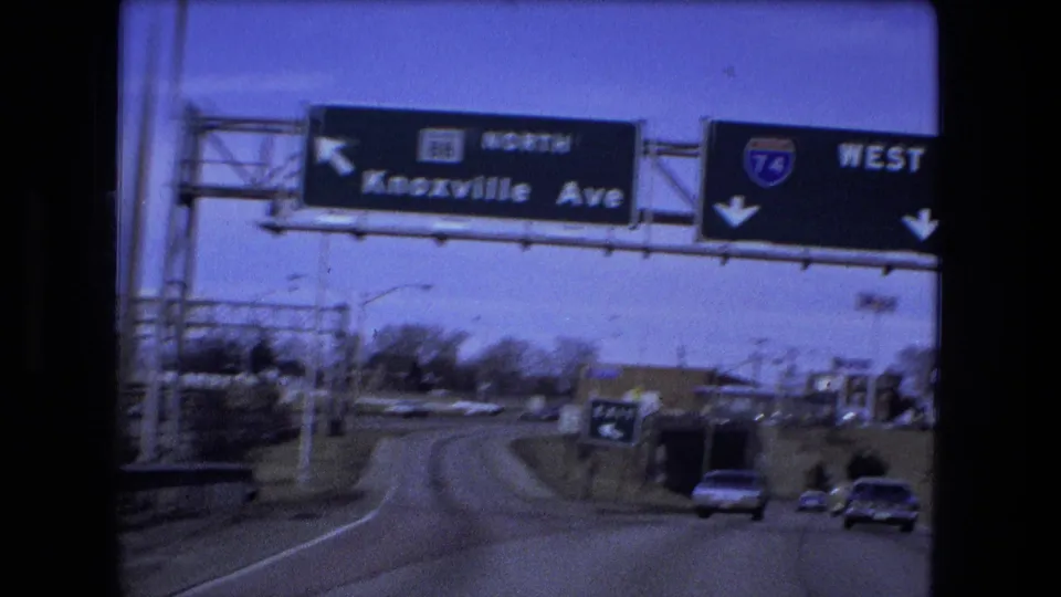 Closer 8mm view of Fort Wayne highway lanes with Knoxville Avenue and I-74 West signs overhead and cars entering an exit area.