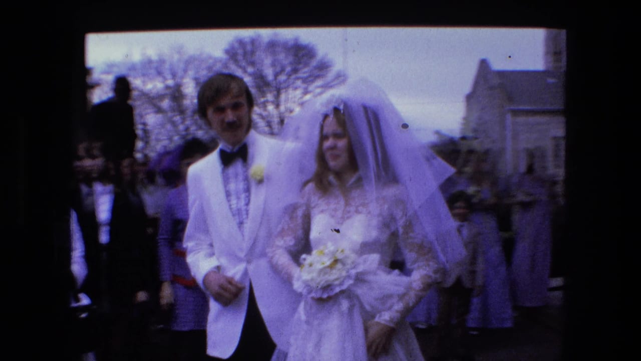 Bride and groom stand close together as guests gather behind them outdoors.