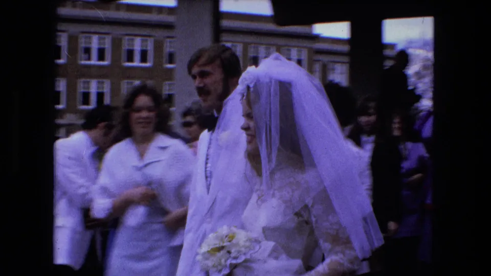 The bride and groom pass close to the camera, with guests and a brick institutional-looking building visible behind them.