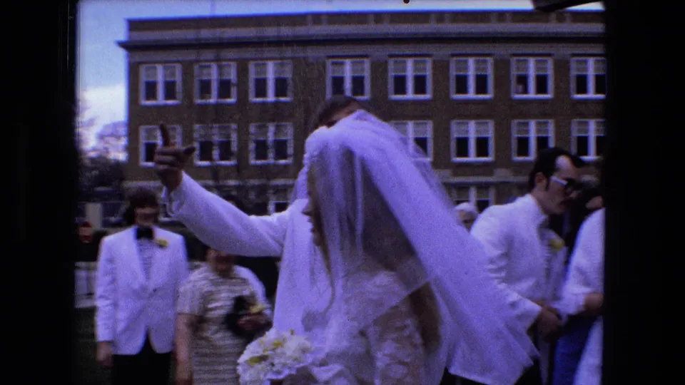 The groom raises an arm as the bride in a long veil moves through guests outside a brick building in Fort Wayne.