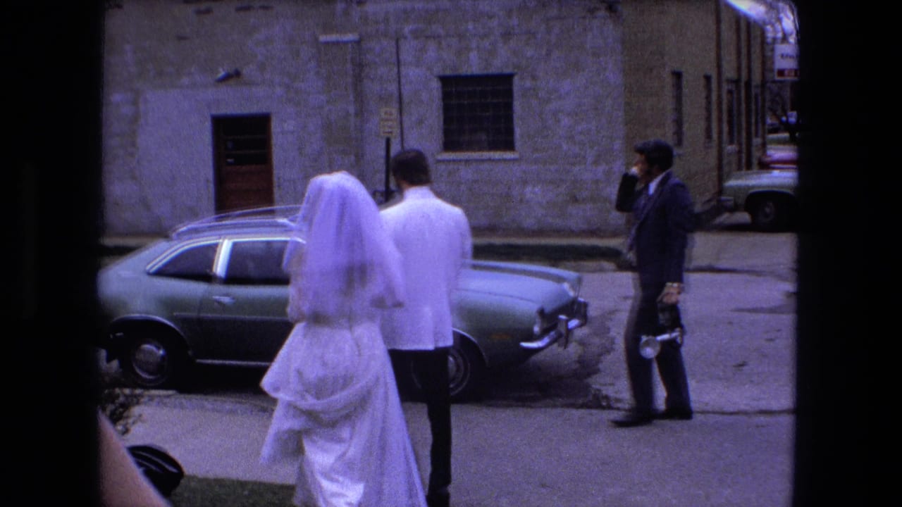 Bride and groom walk along a street toward a parked sedan.