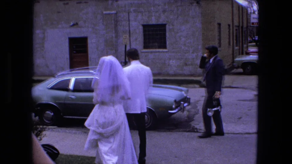 The bride and groom walk on a Fort Wayne street beside a parked green sedan while a photographer stands nearby.