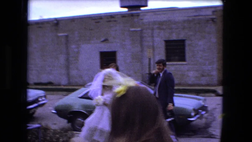 The bride, carrying yellow flowers, moves past guests and parked cars in 1973 Fort Wayne wedding footage.