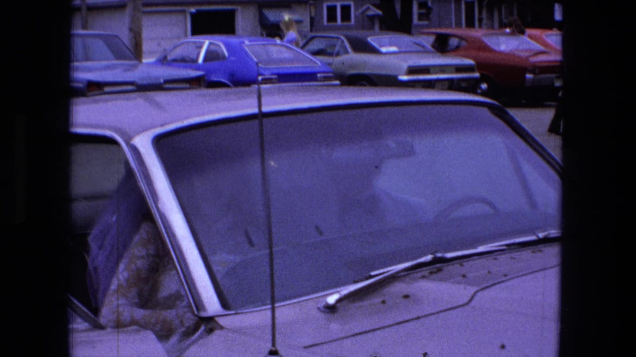 1973 Fort Wayne home-movie frame showing a woman in a wedding dress inside a white car, with period parked cars in the background.