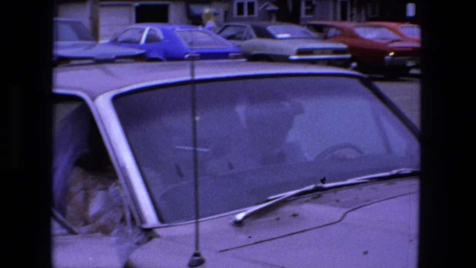 1973 Fort Wayne 8mm frame showing the bride partly obscured inside a white car, with 1970s parked cars beyond.
