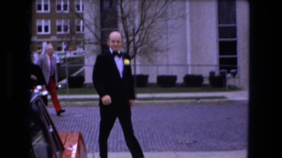 1973 Fort Wayne wedding home-movie frame with a tuxedoed man walking outdoors near cars, buildings, and bare trees.