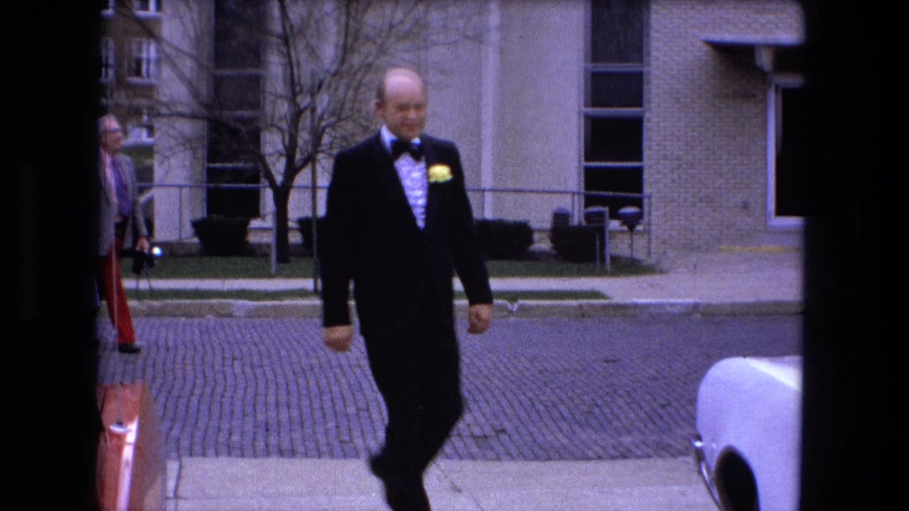 Man in a black tuxedo walks on a sidewalk near a white car and brick building.