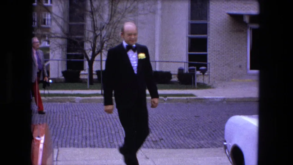 In Fort Wayne, Indiana in 1973, a man in a black tuxedo and yellow boutonniere walks toward a white car outside a brick building.